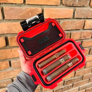 May include: A red YETI waterproof case, open to reveal three cigars. The case has a black interior with a mesh section and a secure latch. The cigars are brown with decorative bands.