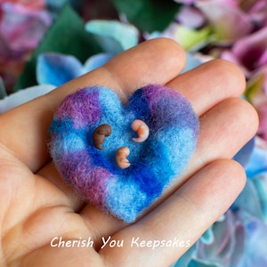 May include: A heart-shaped felt keepsake in shades of blue and purple, with three small, brown and pink objects. The keepsake is held in an open hand, with a blurred floral background. Text at the bottom reads "Cherish You Keepsakes."