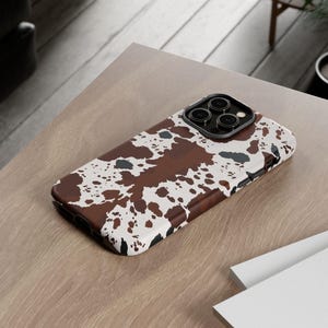 May include: A brown, white, and black cowhide-patterned phone case. The case is lying on a light brown wooden surface. The phone case has a camera cutout and is next to a stack of white books.