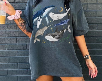 May include: An oversized dark grey t-shirt featuring a graphic print of whales, a narwhal, and a crescent moon. The design is in shades of blue, grey, and white. The shirt has a relaxed fit and a crew neck.