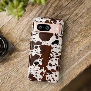 May include: A smartphone case with a cowhide pattern in brown, black, and white. The case is on a wooden surface, with a camera and plant in the background. The phone case has a rose gold camera bar.