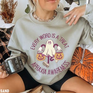Puede incluir: Una sudadera color arena con el texto "EVERY WORD IS A TREAT APRAXIA AWARENESS" que rodea un fantasma de dibujos animados sosteniendo dos calabazas. El diseño es en tonos morado, naranja y blanco.