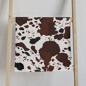 May include: A white towel with a brown and black cowhide pattern is displayed on a wooden ladder. The towel's design features irregular brown and black spots against a white background, creating a rustic aesthetic. The towel is folded and hung on the ladder.