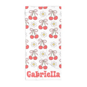 Può includere: Asciugamano da spiaggia bianco con un motivo ripetitivo di fiocchi rosa, ciliegie rosse e margherite bianche. La parte inferiore dell'asciugamano ha il nome "Gabriella" in lettere rosa.