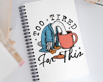 May include: A spiral-bound notebook with a whimsical illustration of a duck wearing a blue robe, with its head in a red mug. The text reads "Too Tired For This" in black script. A brown slipper is also shown.