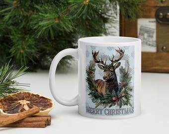 May include: White ceramic mug featuring a detailed illustration of a stag with large antlers, surrounded by greenery and a red bow. The words "MERRY CHRISTMAS" are printed below the image. A festive holiday theme.