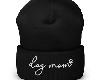 May include: Black beanie with the embroidered text "dog mom" in white script, accompanied by a paw print. The beanie is a solid black color and appears to be made of a soft, knitted material.