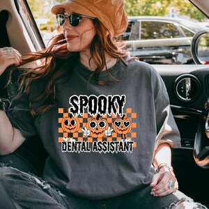 May include: Dark gray t-shirt with the words "SPOOKY DENTAL ASSISTANT" in a black and white font. The shirt features three orange jack-o'-lanterns with different facial expressions on a black and orange checkered background. The shirt is a Halloween-themed item.