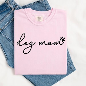 May include: A light pink t-shirt with the words "dog mom" in black script and a paw print. The shirt is folded on top of a pair of blue denim jeans.
