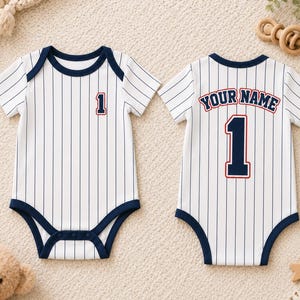May include: Two white baby bodysuits with navy blue pinstripes and navy trim. The front features the number 1 in navy with a red outline. The back displays the text YOUR NAME in navy with a red outline above a large navy number 1 with a red outline.