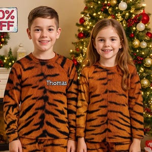 May include: Two children wearing matching tiger-striped pajamas. The pajamas are orange and black, with a long-sleeved top and pants. The boy's pajamas have the name "Thomas" on the chest. A sign in the upper left corner says "50% OFF".