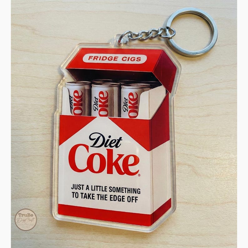 Puede incluir: Un llavero de Diet Coke con forma de paquete de cigarrillos. El llavero presenta el logotipo de Diet Coke y el texto "JUST A LITTLE SOMETHING TO TAKE THE EDGE OFF". La parte superior del paquete dice "FRIDGE CIGS".