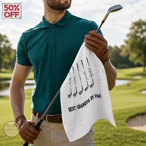 May include: A white golf towel with the text "BEST GRANDPA BY PAR" and golf club illustrations. The towel also features names. The image shows a person holding the towel and a golf club. A "50% OFF" sticker is in the top left corner.
