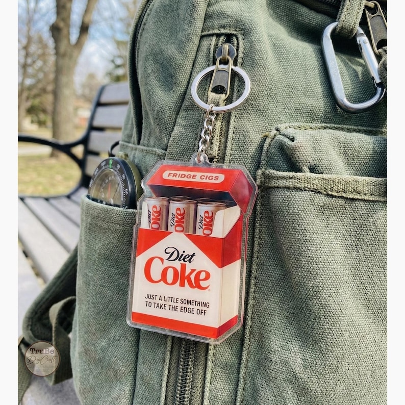 Puede incluir: Un llavero novedoso con un dise&ntilde;o de paquete de cigarrillos Diet Coke en miniatura. El paquete rojo y blanco dice "Diet Coke" y "Fridge Cigs" con el lema "Just a little something to take the edge off."