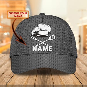 May include: A gray baseball cap with a honeycomb pattern. The cap features a white chef's hat, crossed spatula and fork, and the word "NAME" in white. The text "CUSTOM YOUR NAME" is in a yellow box.