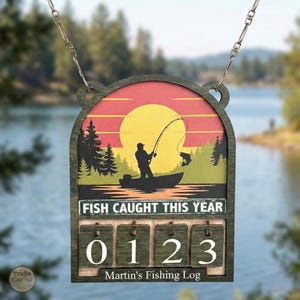 Puede incluir: Registro de pesca de madera con diseño de puesta de sol. Presenta la silueta de un pescador en un bote, pescando un pez. El texto "FISH CAUGHT THIS YEAR" está sobre los diales numéricos, actualmente en 0123. Colgando de una cadena.