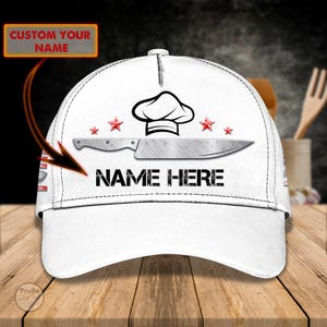 May include: White baseball cap featuring a chef's hat, knife, and red star graphics. The text "NAME HERE" is printed below the knife. The top left corner has the text "CUSTOM YOUR NAME" in a rectangular box.