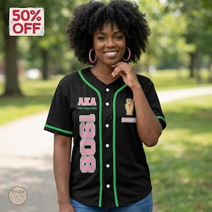 Op de afbeelding: Zwart honkbalshirt met groene en witte afwerking. Het shirt toont de letters "AKA" en het nummer "1908" in roze. Het heeft ook een vuistafbeelding en de naam "Jackson".