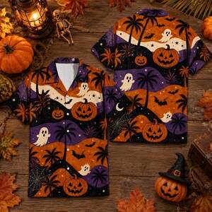 May include: A short-sleeve button-down shirt featuring a Halloween-themed pattern with orange pumpkins, white ghosts, black bats, and palm trees against wavy bands of orange, purple, and black. The shirt has a notched collar and a button-front closure.
