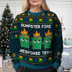 Könnte beinhalten: Ein teal-farbener Weihnachtspullover mit dem weißen Schriftzug "DUMPSTER FIRE RESPONSE TEAM". Der Pullover zeigt drei grüne Cartoon-Mülltonnen mit Flammen, die jeweils mit einem Namen versehen sind. Das Design umfasst Rentiere, Schneeflocken und Weihnachtsbäume.