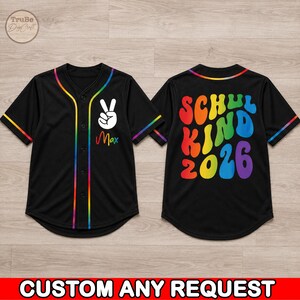 May include: A black baseball jersey with rainbow trim along the collar, sleeves, and front placket. The front features a white peace sign hand gesture and the name Max. The back displays the colorful text SCHUL KIND 2026 in a wavy font.