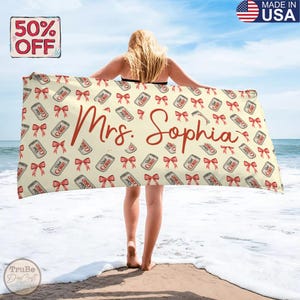 Puede incluir: Una toalla de playa personalizada con un fondo crema, con un patrón de latas de Diet Coke y lazos rojos. El nombre "Mrs. Sophia" está escrito en rojo. La toalla está en una playa.
