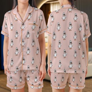 Peut inclure: Ensemble de pyjama rose avec un motif répété de canards blancs portant des lunettes de soleil. Le haut à manches courtes et le short ont une bordure orange. L'ensemble est en matière douce et soyeuse.