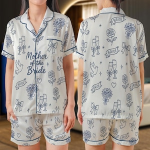 May include: A white pajama set with a navy blue celebratory print. The short-sleeved top and shorts feature a pattern of doves, roses, bows, champagne glasses, and the text "Mother of the Bride" and "Bride".