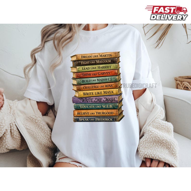 May include: White t-shirt featuring a stack of books with inspirational phrases like "Dream Like Martin" and "Write Like Maya." The books have a vintage look with gold lettering and are stacked on top of each other. The shirt is a casual style.