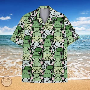 May include: A short-sleeved button-down shirt featuring a repeating pattern of cartoon frogs in shades of green, pink, and white. The shirt has a classic collar and is set against a beach backdrop with blue skies and ocean.
