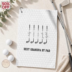 May include: White golf towel with a textured pattern, featuring black golf club illustrations with names and the text "BEST GRANDPA BY PAR." Includes golf balls and clubs.