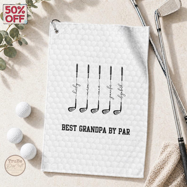 May include: White golf towel with a textured pattern, featuring black golf club illustrations with names and the text "BEST GRANDPA BY PAR." Includes golf balls and clubs.