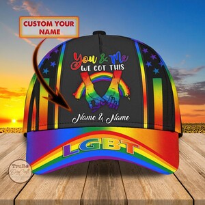 Op de afbeelding: Zwarte baseballcap met een regenboogrand en accenten. De cap heeft de tekst "You & Me WE GOT THIS" en "LGBT". Het ontwerp bevat een regenboog en twee handen die elkaar vasthouden. De tekst "Name & Name" is ook zichtbaar.