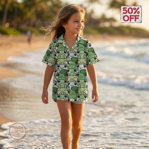 Pode incluir: Uma camisa verde de manga curta com um padrão repetido de rostos de sapos. A camisa é usada por uma pessoa na praia. Uma placa no canto superior direito diz "50% OFF".