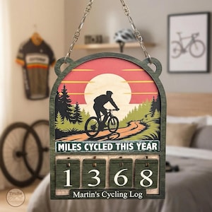 May include: Wooden cycling log with a green frame and chain. The artwork depicts a cyclist on a winding path with a sunset backdrop. The log displays "MILES CYCLED THIS YEAR" and the number "1368". The text "Martin's Cycling Log" is at the bottom.