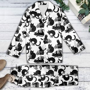 Pode incluir: Conjunto de pijama branco com padrão de gatos pretos. A parte superior de manga comprida tem uma frente abotoada e uma gola entalhada. As calças a condizer têm uma cintura elástica. Os gatos estão em várias poses, alguns tocando instrumentos.