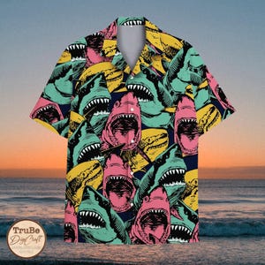 May include: A short-sleeved button-up shirt with a colourful print of sharks with open mouths. The shirt is on a background of a beach with ocean waves and a sunset sky.