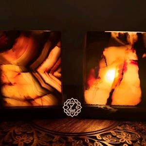 May include: Two rectangular stone lamps with internal illumination, displaying warm orange, amber, and dark brown veined patterns. The left lamp features sharp geometric banding, while the right lamp shows organic, flowing mineral inclusions.
