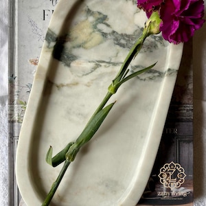 May include: An oval marble tray with green and gray veining, holding a vibrant purple carnation. The tray rests on a book with the text "the F" visible. The Etsy shop name "ZanyLiving" is also visible.