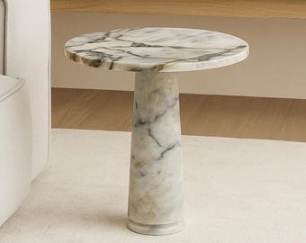 Puede incluir: Una mesa auxiliar redonda de mármol con una tapa con diseño blanco y gris. La mesa tiene una base cilíndrica y se apoya sobre una alfombra blanca. Es un mueble elegante.
