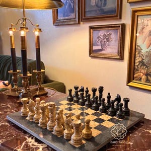 May include: A chess set with black and beige pieces on a checkered board. The board is set on a marble-like surface. A decorative lamp and framed artwork are in the background. The chess set is a classic game.