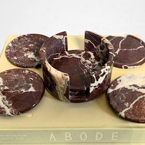 May include: A set of marble coasters and a matching holder. The coasters and holder are a deep burgundy color with white veining. The set is displayed on a book with the word "ABODE" visible.