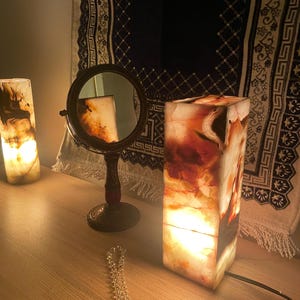 May include: A warm, inviting scene featuring two illuminated onyx lamps with a marbled appearance, a small round mirror, and a decorative beaded necklace. The lamps emit a soft glow, creating a cozy ambiance. The lamps are rectangular and the mirror is round.