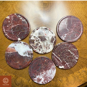 May include: Six round coasters with a polished finish. The coasters are made of a reddish-brown stone with white and gray veining. The coasters are arranged on a wooden surface. The logo "zany living" is in the bottom left corner.