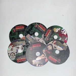 May include: A selection of six DVDs from the anime series "Baki the Grappler." The discs are circular, displaying character illustrations and the series title. Each disc is numbered, with visible numbers 7, 8, 9, 11, and 12.