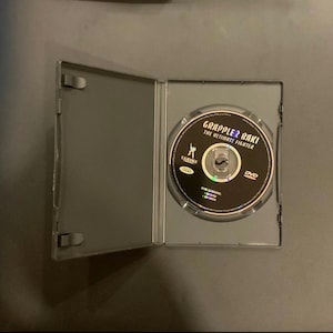 Puede incluir: Una caja de DVD negra abierta revela un disco con la etiqueta "Grappler Baki: The Ultimate Fighter". El disco tiene un fondo negro con texto blanco y dorado. La caja es gris y est&aacute; sobre una superficie oscura.