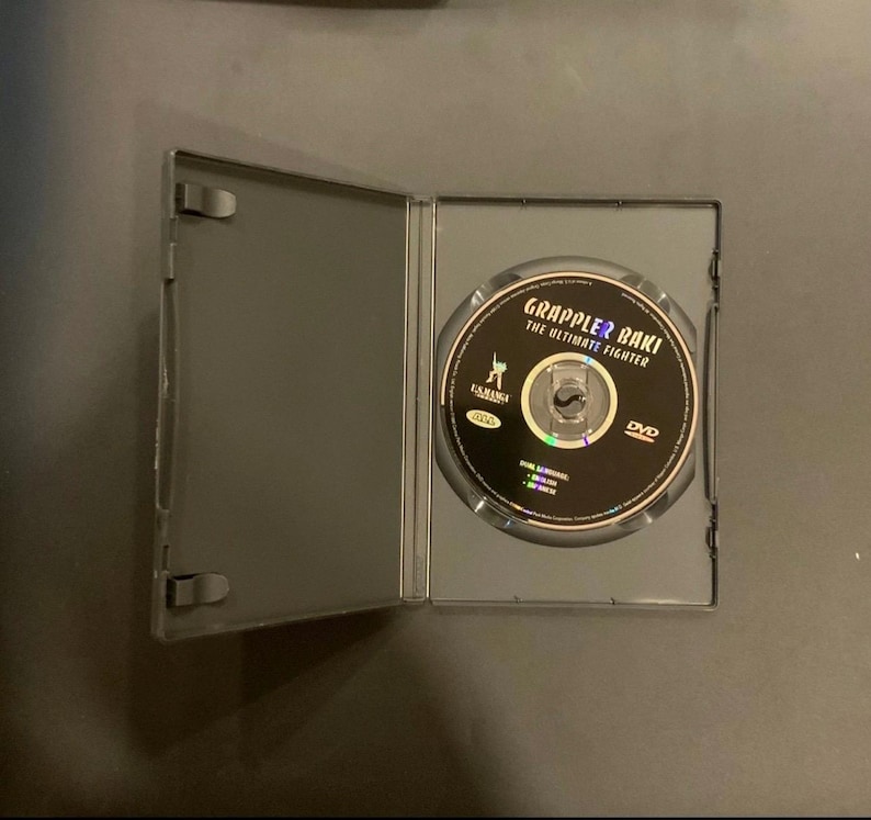 Puede incluir: Una caja de DVD negra abierta revela un disco con la etiqueta "Grappler Baki: The Ultimate Fighter". El disco tiene un fondo negro con texto blanco y dorado. La caja es gris y est&aacute; sobre una superficie oscura.