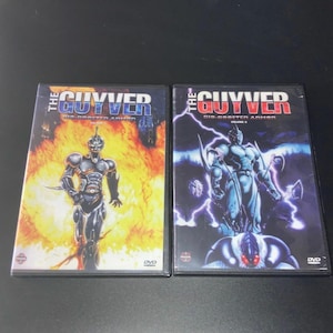 Puede incluir: Dos estuches de DVD de "The Guyver: Bio-Booster Armor". Un estuche presenta un personaje con armadura plateada sobre un fondo de fuego. El otro estuche muestra un personaje similar con un efecto de relámpagos azules y morados.