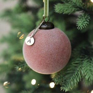May include: A dusty rose Christmas ornament with a velvety texture hangs from a green ribbon. A small, round gold charm with the letter "A" dangles from the ornament. The ornament is set against a backdrop of a green Christmas tree with soft, glowing lights.