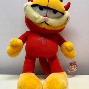 May include: Red plush Garfield toy with devil horns and a red and black striped tail. The toy is wearing a red shirt with the words "Hot Stuff" printed on it. The toy has a yellow face with black eyes and a pink nose.